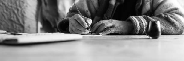 Wide low angle view of an elderly man doing calligraphy writing using a nib pen and ink on a sheet of white paper in a monochrome image.